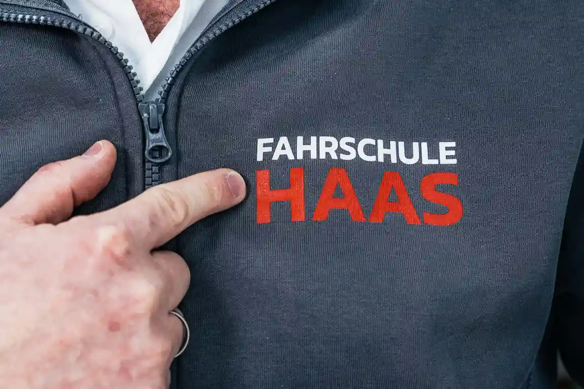 Fahrschullehrer und Ausbilder für Berufskraftfahrer zeigt mit seinem Finger auf das Logo der Fahrschule auf seinem Pullover.